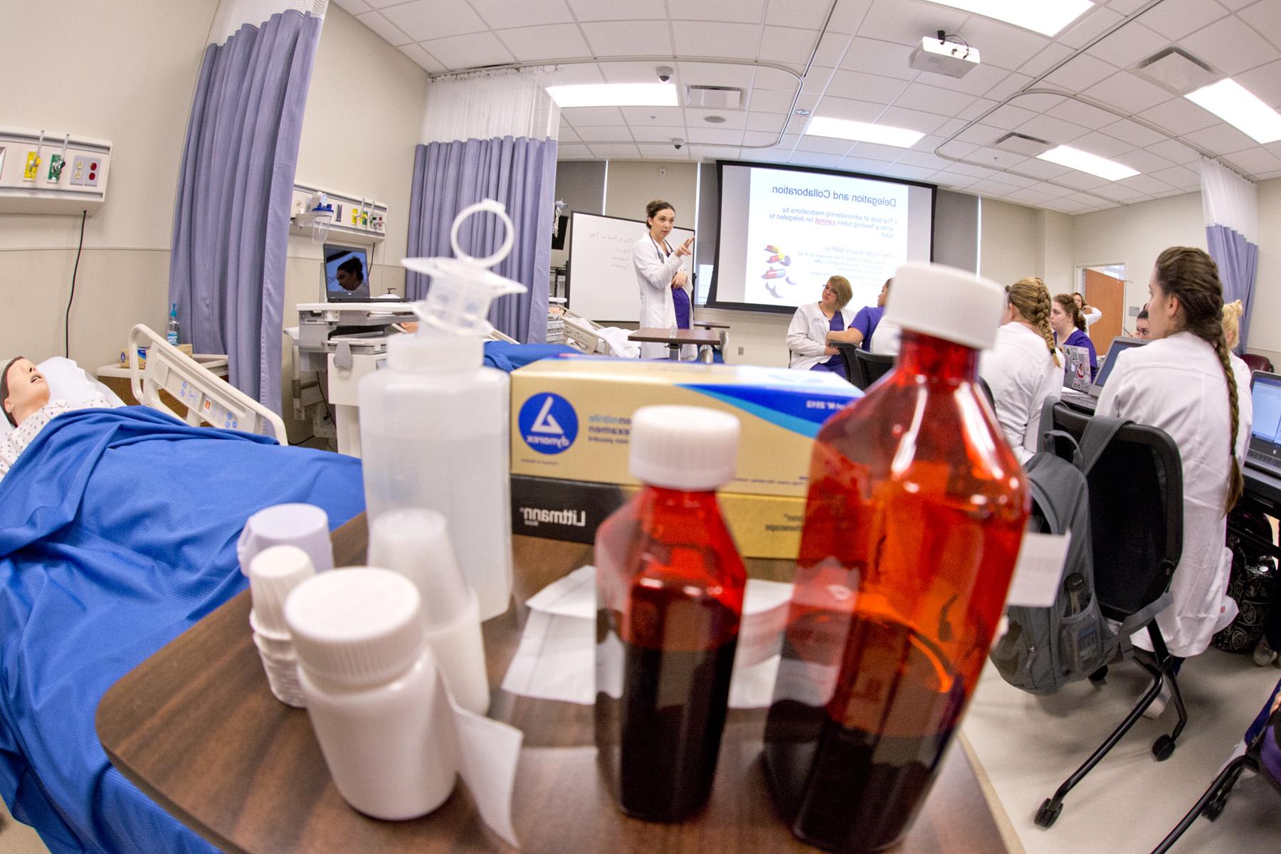 Students in white coats listen to a female professor in front of a projector in a nursing simulation lab surrounded by medical equipment and prescription bottles